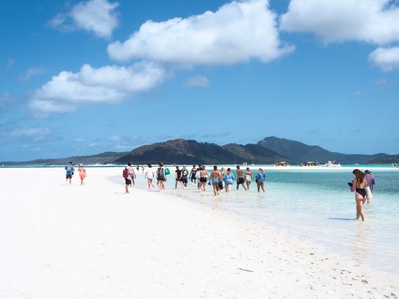 fotografía de un grupo de jóvenes de espalda que anda por la playa de las islas Whitsundays. Al fondo las montañas